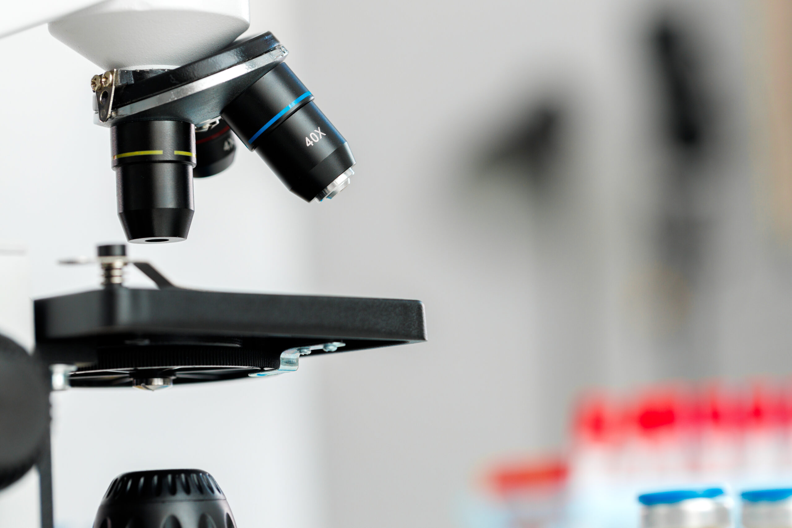 Detal mikroskopu optycznego z widocznym obiektywem o powiększeniu 40x. W tle widoczne rozmyte fiolki laboratoryjne z czerwonymi nakrętkami, sugerujące środowisko badawcze lub medyczne.